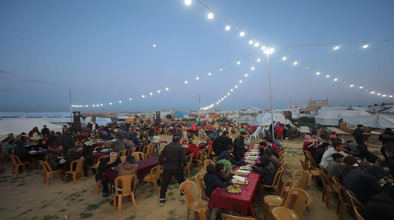 مواطنون يتناولون إفطارهم خلال شهر رمضان ضمن برنامج إفطار جماعي نظمته منظمة خيرية للنازحين، وسط الأنقاض في مخيم البريج بوسط قطاع غزة